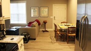 A compact kitchen with a refrigerator, stove, and coffee maker. There is a dining table with chairs and a sofa with a red blanket. Two framed pictures are on the wall, and a door is visible in the background. - Haley Retreat BELLA VISTA LOCATION/BENTONVILLE ADDRESS.ONE MILE TO OZ BIKE LIFT. (Bentonville)