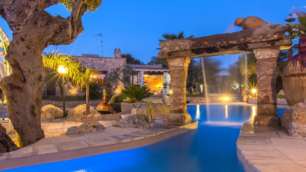 Un área de piscina con una estructura de piedra, un árbol y un edificio al fondo.