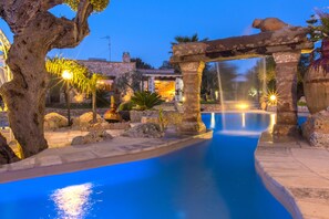 A pool area with a stone structure, a tree, and a building in the background. - Beautiful villa for 12 people with private pool and panoramic view (castrignano del capo)