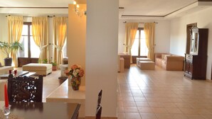 Un vaste salon comprenant un canapé, des fauteuils, une table basse et une table à manger avec des chaises. Les fenêtres sont dotées de rideaux, et on y trouve un lustre ainsi qu’un meuble avec un miroir.