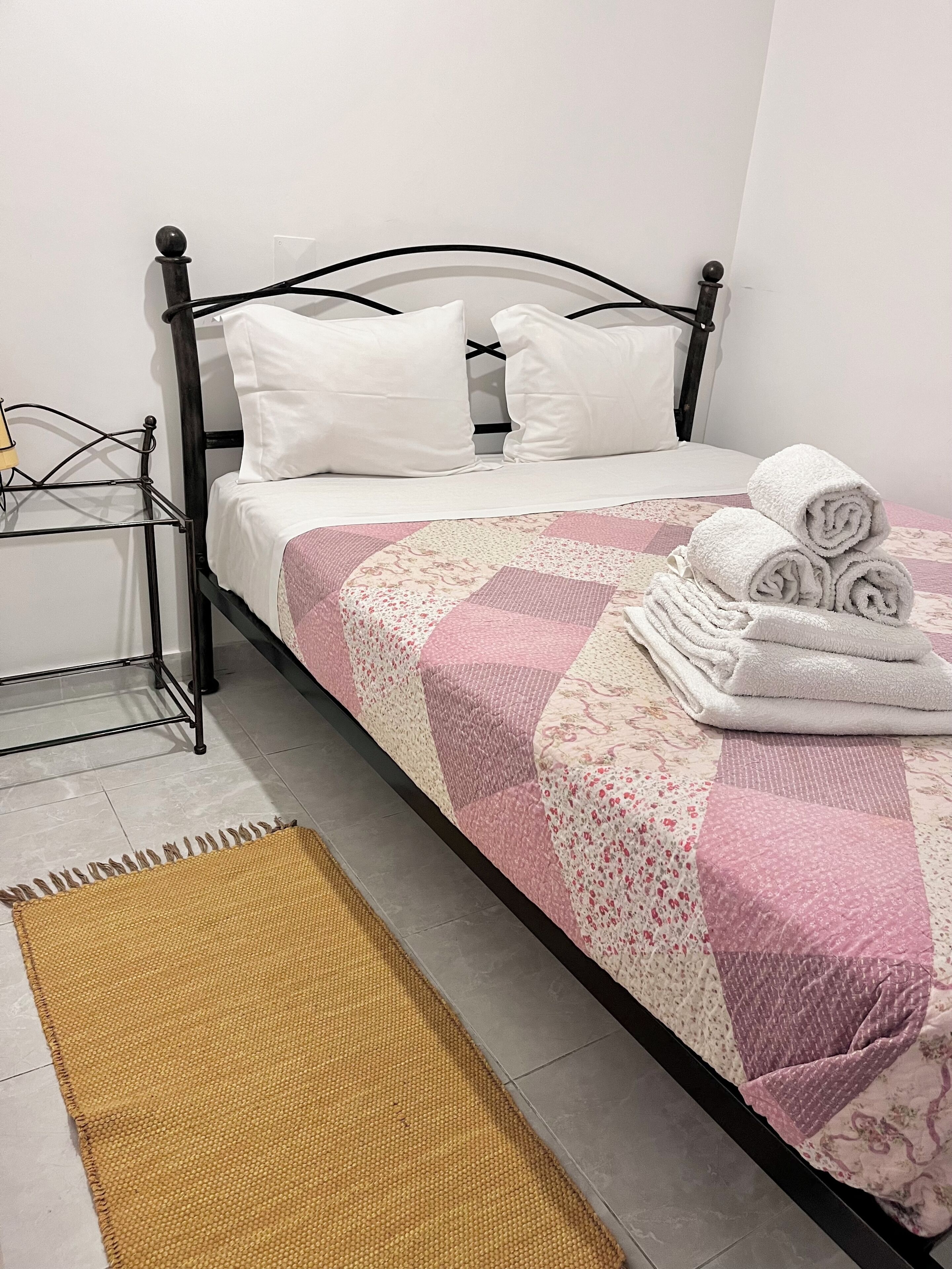 A bed with white pillows, a pink and white quilt, and rolled towels on the side.