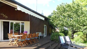 Une terrasse en bois avec des meubles d’extérieur, un patio couvert et une vue sur un jardin agrémenté d’un grand arbre.