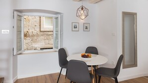 Un coin repas avec une table ronde, quatre chaises, un panier de fruits, un luminaire suspendu, un mur en pierre et une porte.