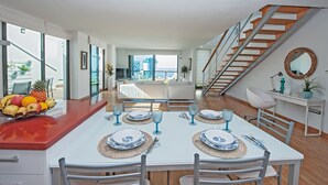 Un espace repas moderne avec une table blanche dressée pour six personnes, un panier de fruits sur le comptoir et un escalier menant à un niveau supérieur.