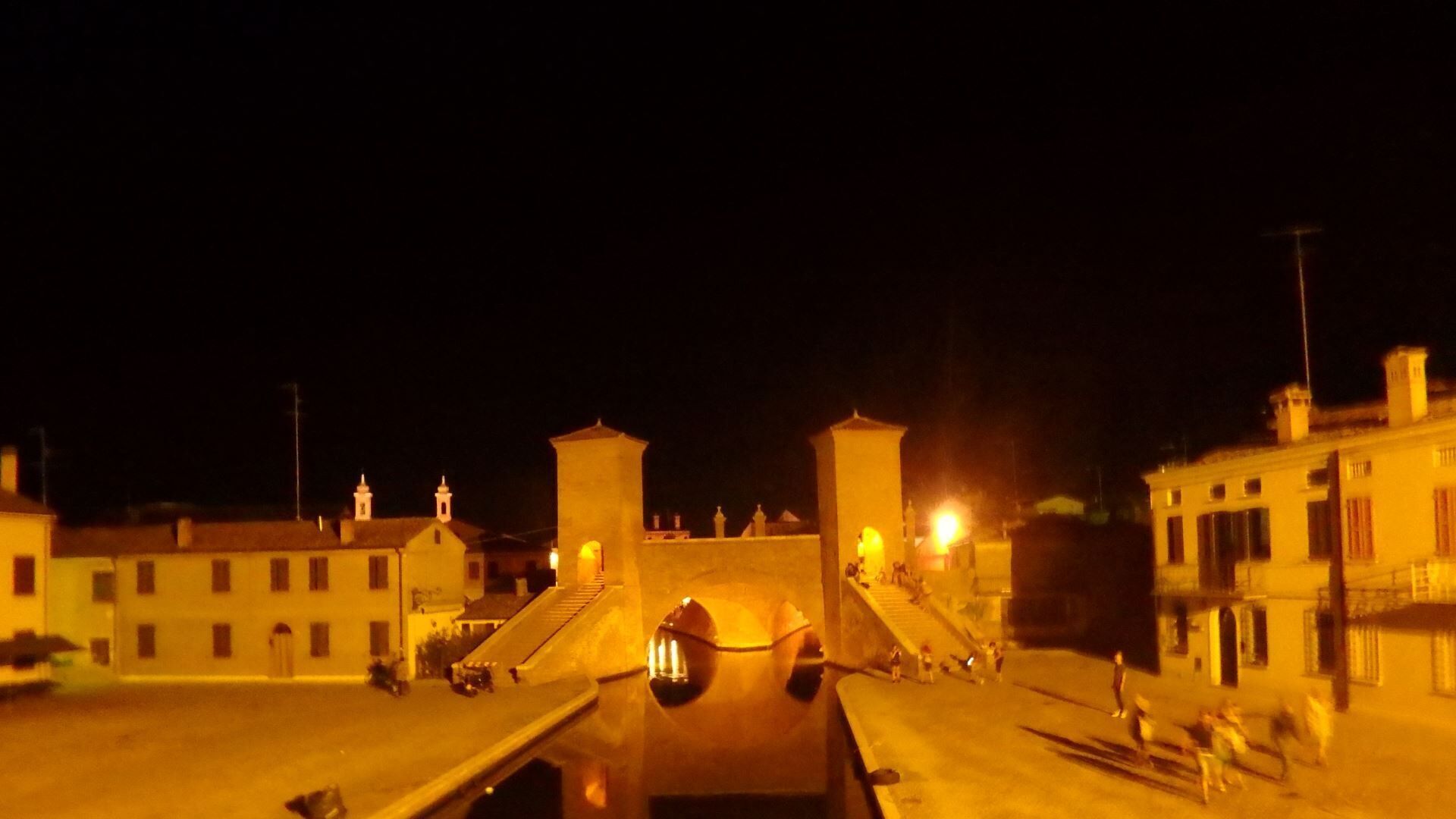 A nighttime view of a waterfront area with buildings, a bridge, and a small boat docked in the water.