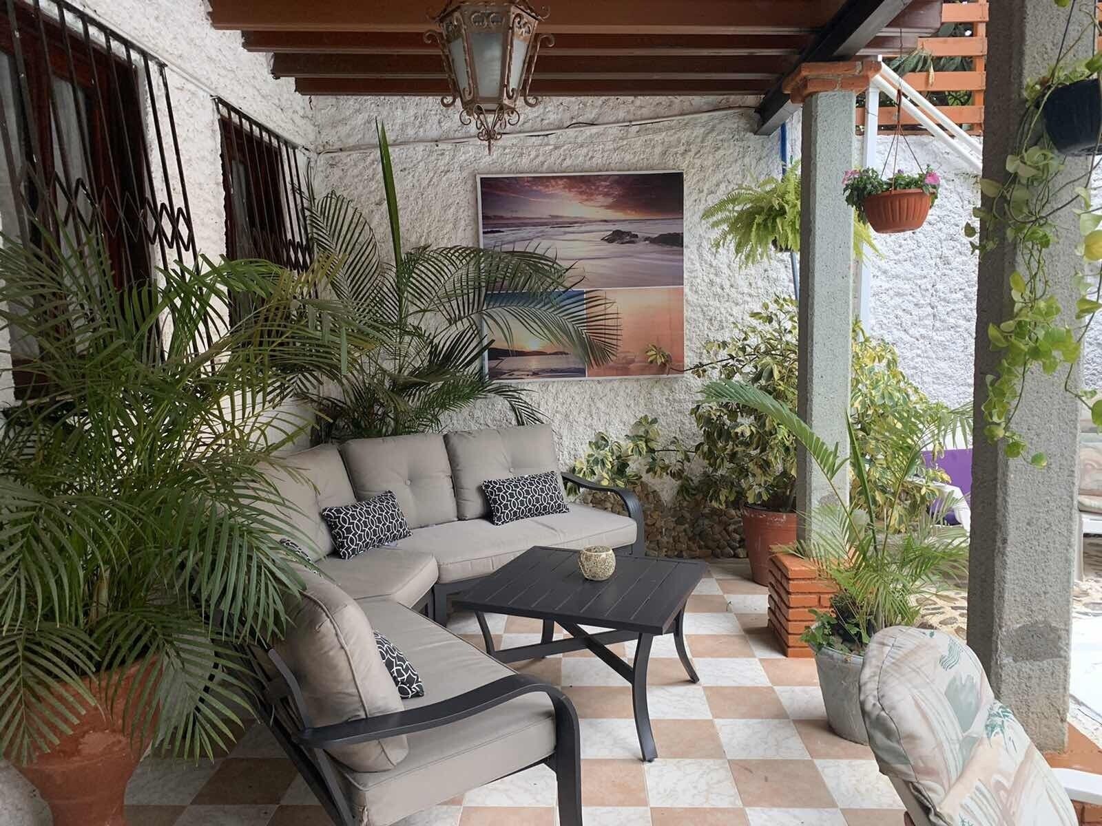 Un área de estar acogedora al aire libre, con un sofá y una mesa de centro, rodeada de plantas en macetas y un cuadro de paisaje.