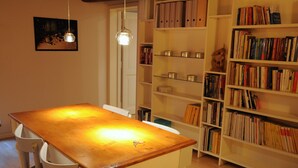 Uma área de refeições com mesa e cadeiras de madeira, uma estante com livros e um lustre.
