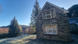 A stone house with two windows, surrounded by a forested landscape and a clear sky.
