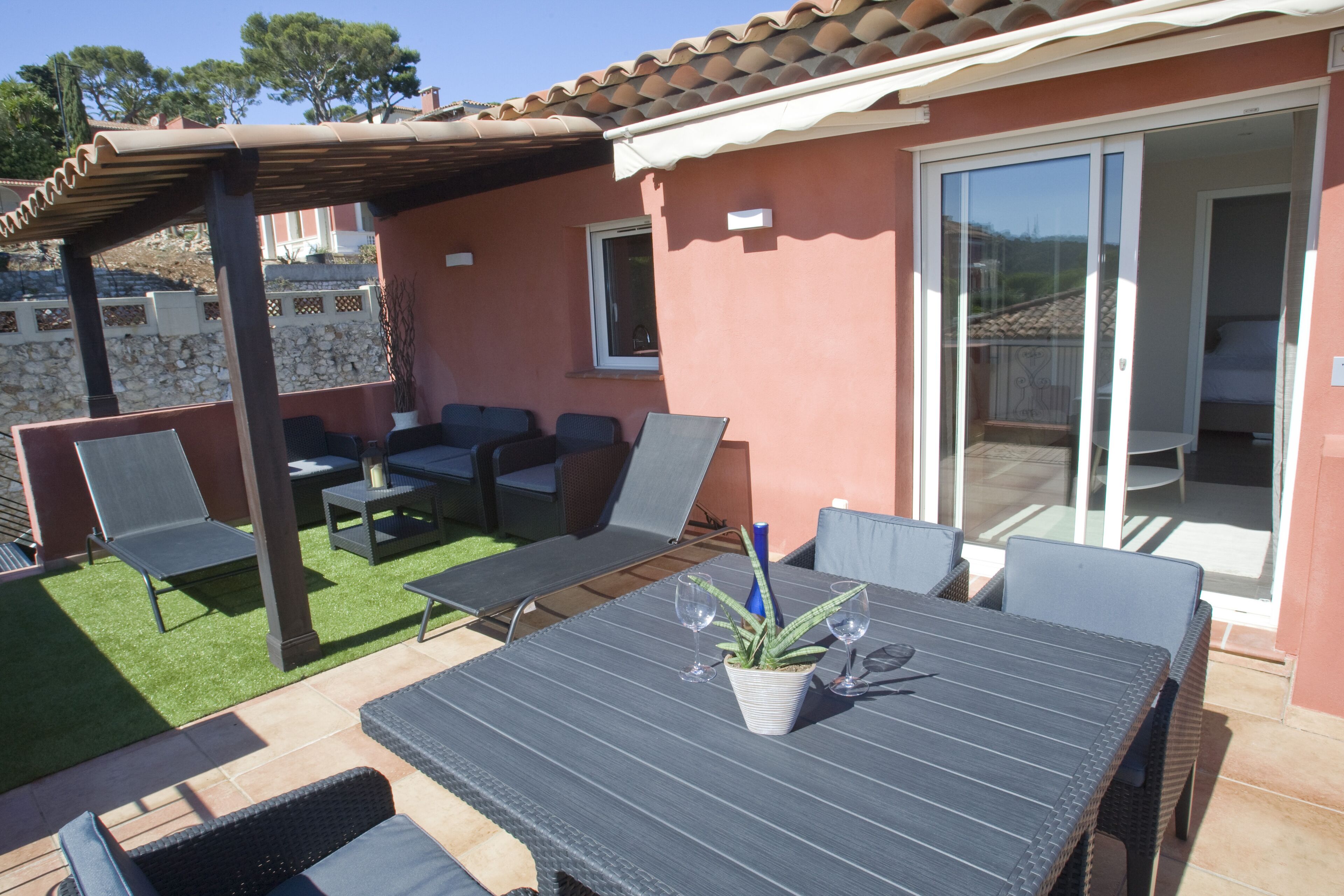 En udendørs terrasse med et bord, stole og et pergolaoverdækket område.