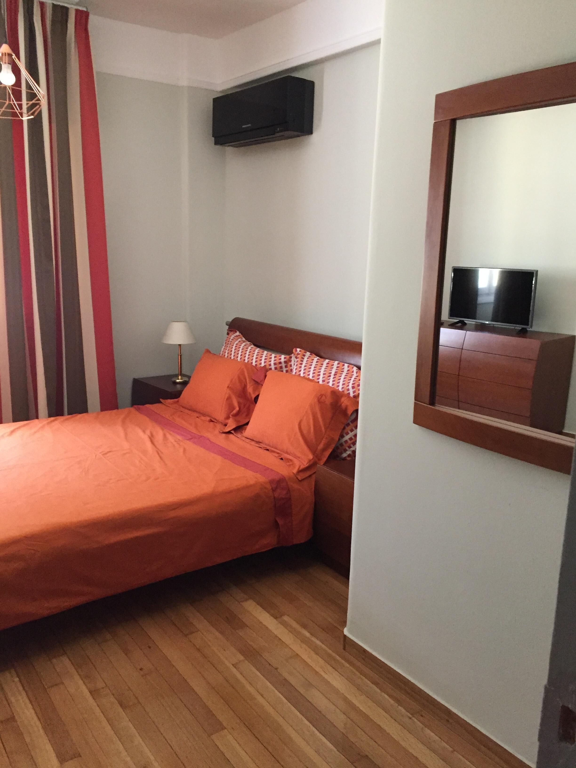 Un dormitorio con cama, mesita de noche, lámpara, televisor sobre un mueble bajo y ventana con cortinas.