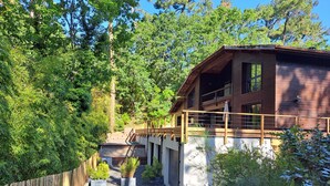 Une cabane en bois dotée d’un balcon, entourée d’une végétation luxuriante et accessible par un chemin de gravier menant à l’entrée.