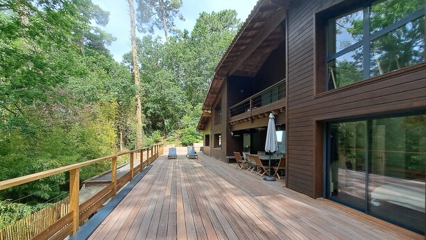 Une terrasse en bois avec une rambarde, un espace repas avec des tables et des chaises, et une grande fenêtre donnant sur une zone boisée.