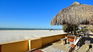Eine Strandterrasse mit einem Reetdachschirm, Holzliegen und Blick auf Strand und Meer.