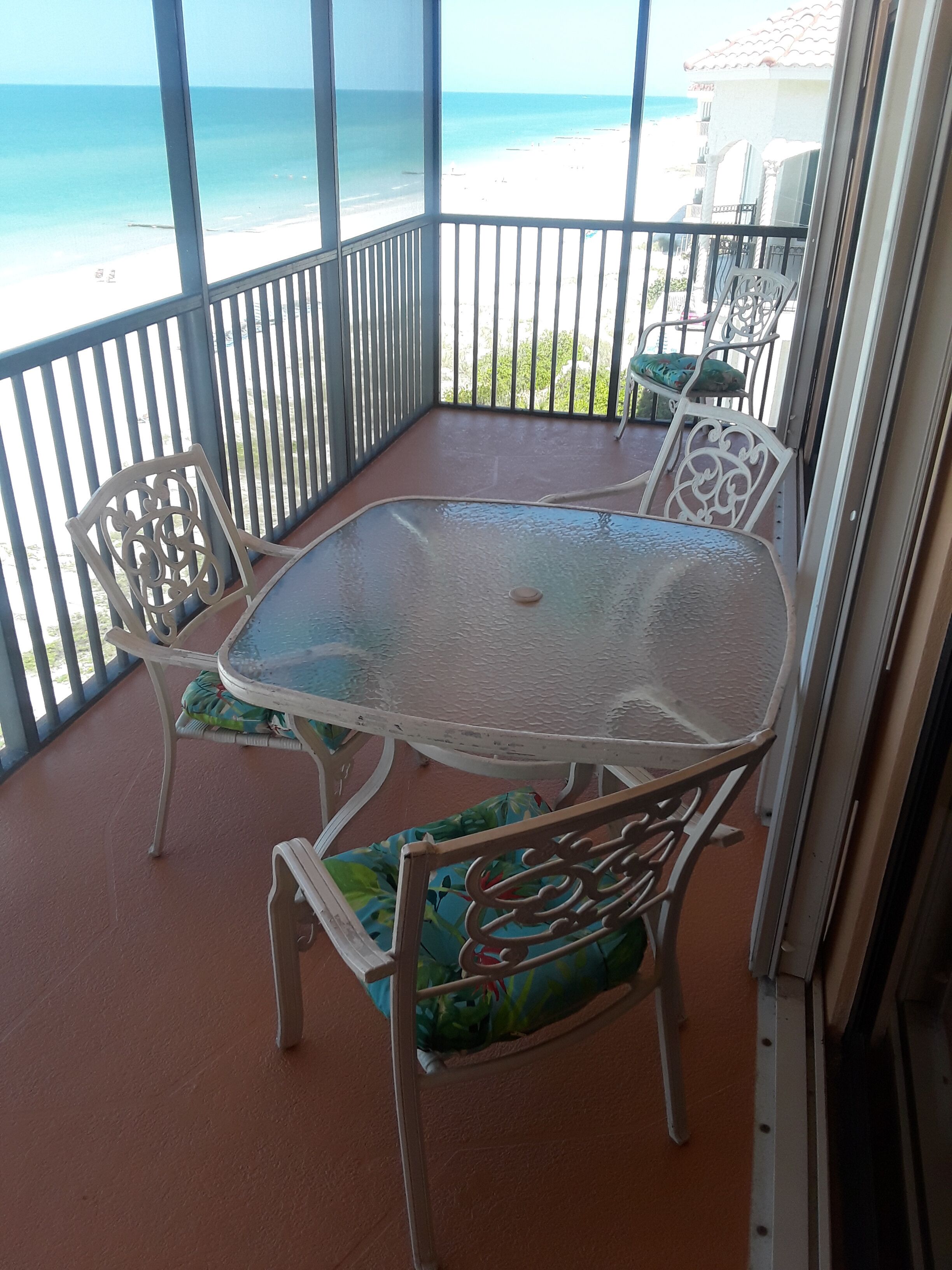 Mesa con cubierta de vidrio y sillas en un balcón con vista a la playa.
