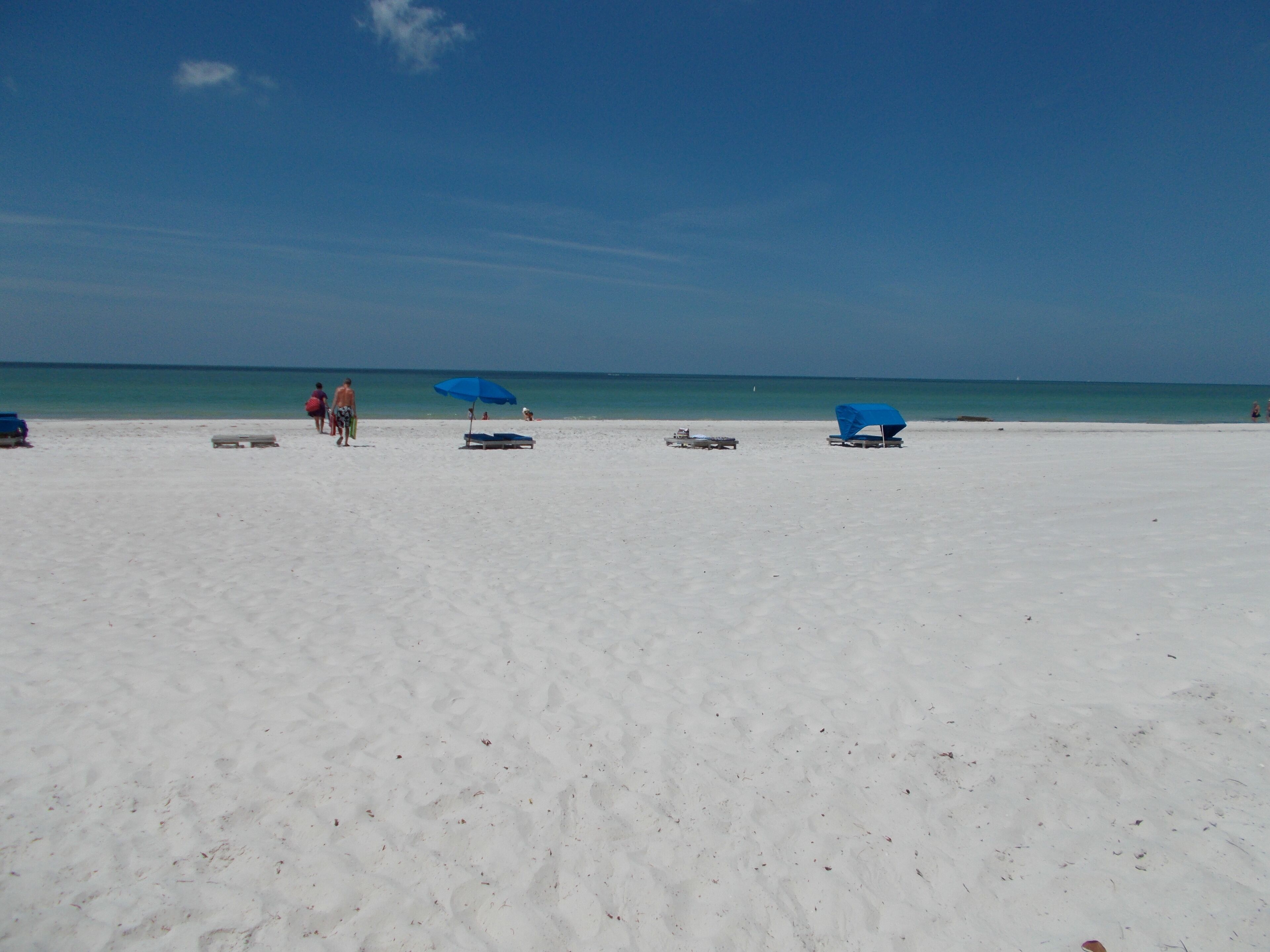 Ein Strand mit weißem Sand und blauem Himmel ohne Wolken. Einige Personen und ein blauer Sonnenschirm sind zu sehen.