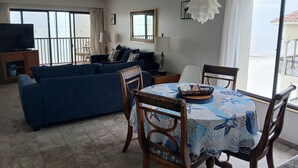 Ein Wohnzimmer mit blauem Sofa, einem Esstisch mit Stühlen, einem Fernseher auf einer Kommode und großen Fenstern mit Meerblick.