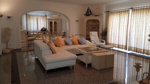 Ein geräumiges Wohnzimmer mit einem weißen Sofa, einem Glastisch und einem weißen Sessel. Im Hintergrund befindet sich eine Essecke mit Tisch und Stühlen.