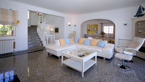 Ein modernes Wohnzimmer mit einem weißen Sofa, einem Glastisch und einem weißen Sessel. Eine Treppe führt auf eine obere Ebene, und im Hintergrund ist ein Essbereich zu sehen.