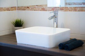 Un bagno con un lavandino bianco, un rubinetto moderno, una piccola pianta in vaso e un asciugamano arrotolato sul ripiano.