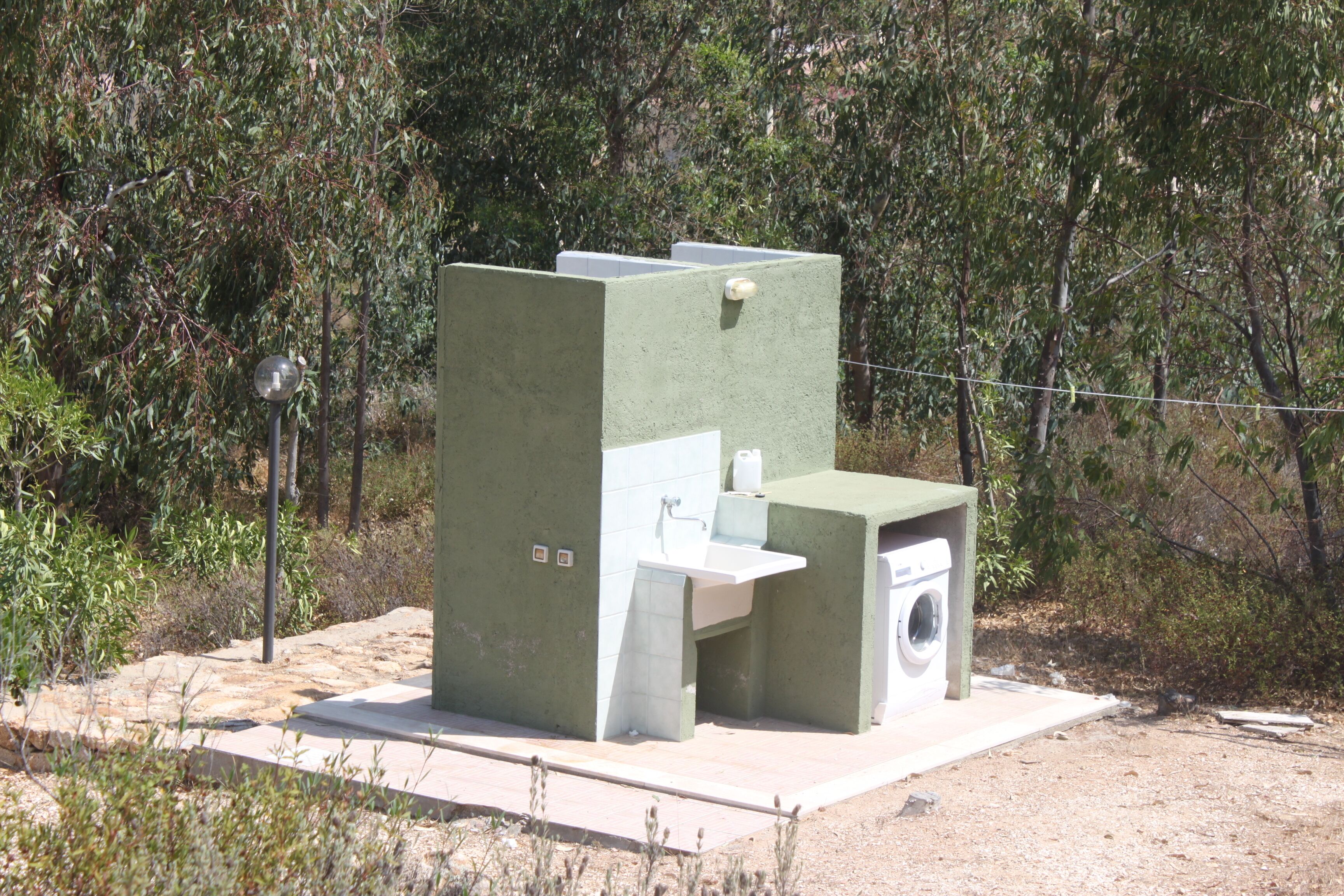 A portable outdoor toilet with a sink and a washing machine, surrounded by natural vegetation.
