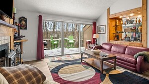 Un salon chaleureux avec une cheminée, un canapé en cuir et une grande fenêtre donnant sur une terrasse où se trouvent des chaises vertes.
