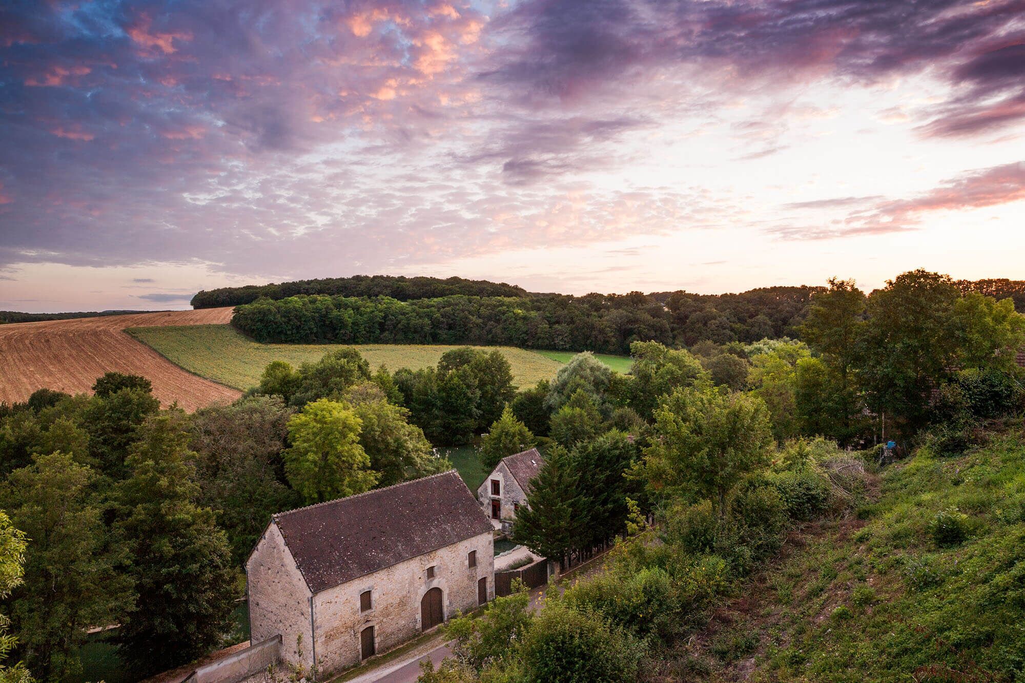 A rural landscape with a stone building surrounded by trees and fields.
