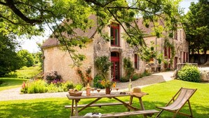 Un edificio rústico de piedra con puerta roja, rodeado de abundante vegetación y un espacio para picnic con banco y mesa.
