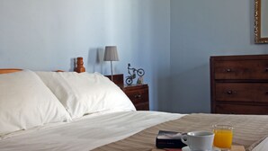 Ein ordentlich bezogenes Bett mit weißen Leinentüchern, ein Nachttisch mit einer Lampe, ein kleines dekoratives Objekt und ein Glas Orangensaft auf einem Tablett.