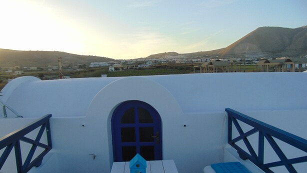A white building with a blue door and windows, a white table with a blue birdhouse, and blue cushions on white benches. - Sunny with sea view apartment (Santorini)