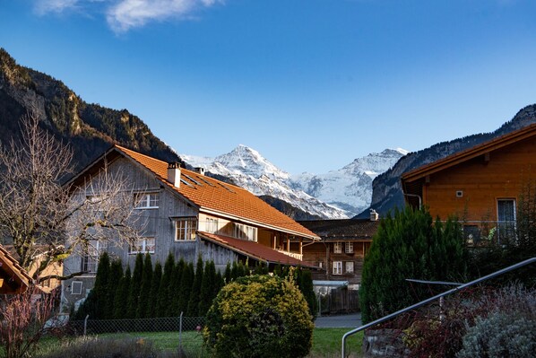 A wooden chalet with a red roof, surrounded by a fenced yard and evergreen trees, with snow-capped mountains in the background.