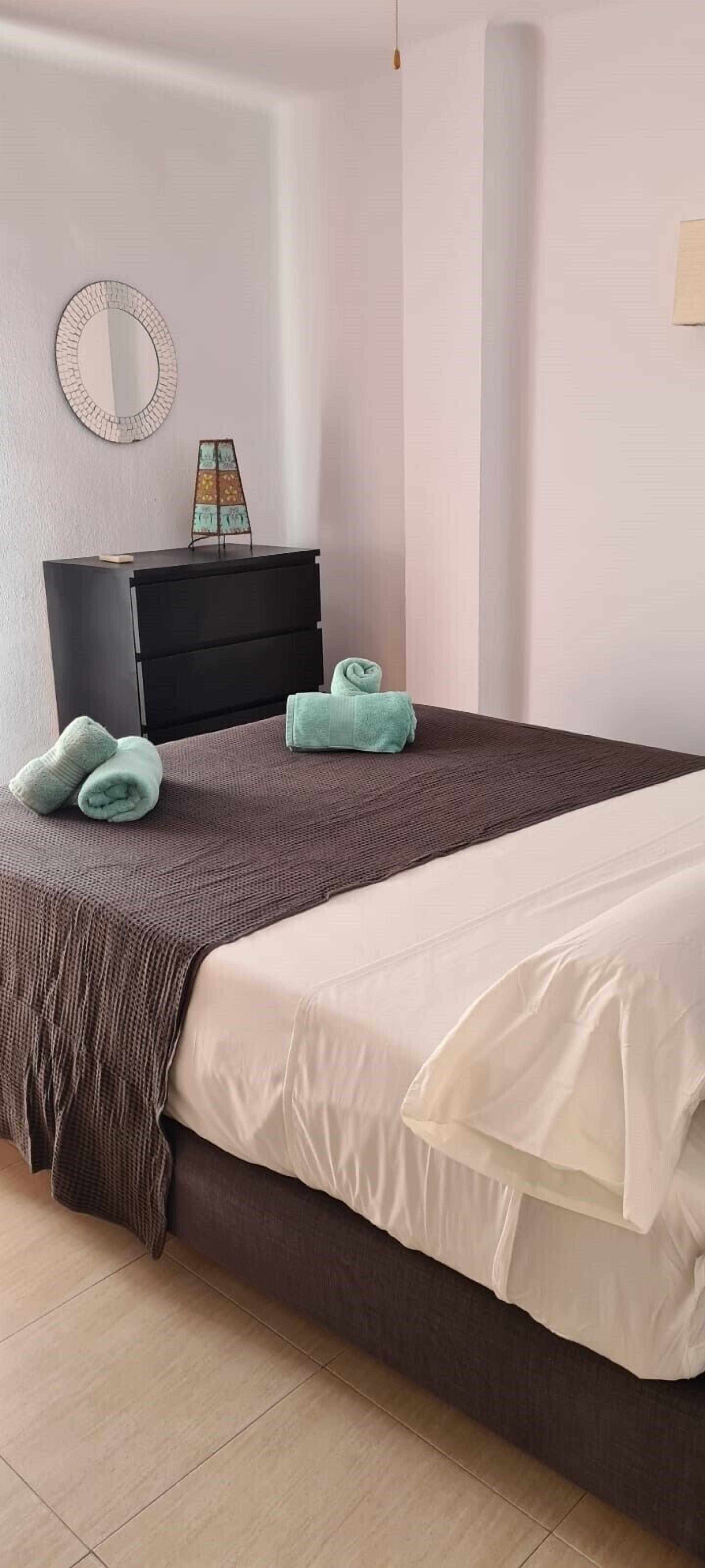 A neatly made bed with a brown and white duvet, a black dresser with a mirror and a small decorative item, and a light-colored wall.