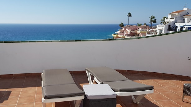 Ein Balkon mit zwei Liegestühlen und einem kleinen Tisch, mit Blick auf die Küste, Gebäude und Palmen.