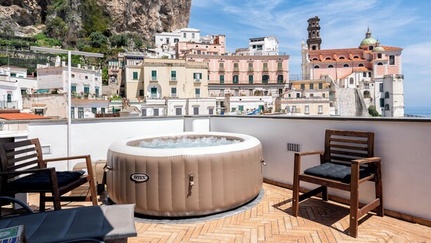 Eine Dachterrasse mit Whirlpool, Holzstühlen und Blick auf historische Gebäude und eine Klippe.