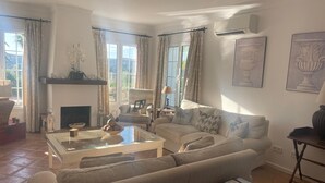 A spacious living room with a large sofa, a glass coffee table, and a fireplace. There are two windows with curtains, and an air conditioning unit is mounted on the wall. - 3 bedrooms, 3 bathrooms, private heated pool, garden area and golf course views (San Roque)
