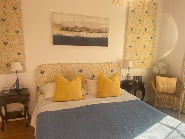 A bedroom with a bed, two bedside tables, a chair, a wall-mounted fan, and a painting.