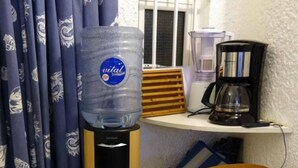 A water dispenser with a bottle of water, a coffee maker, and a chair in a room with patterned curtains.