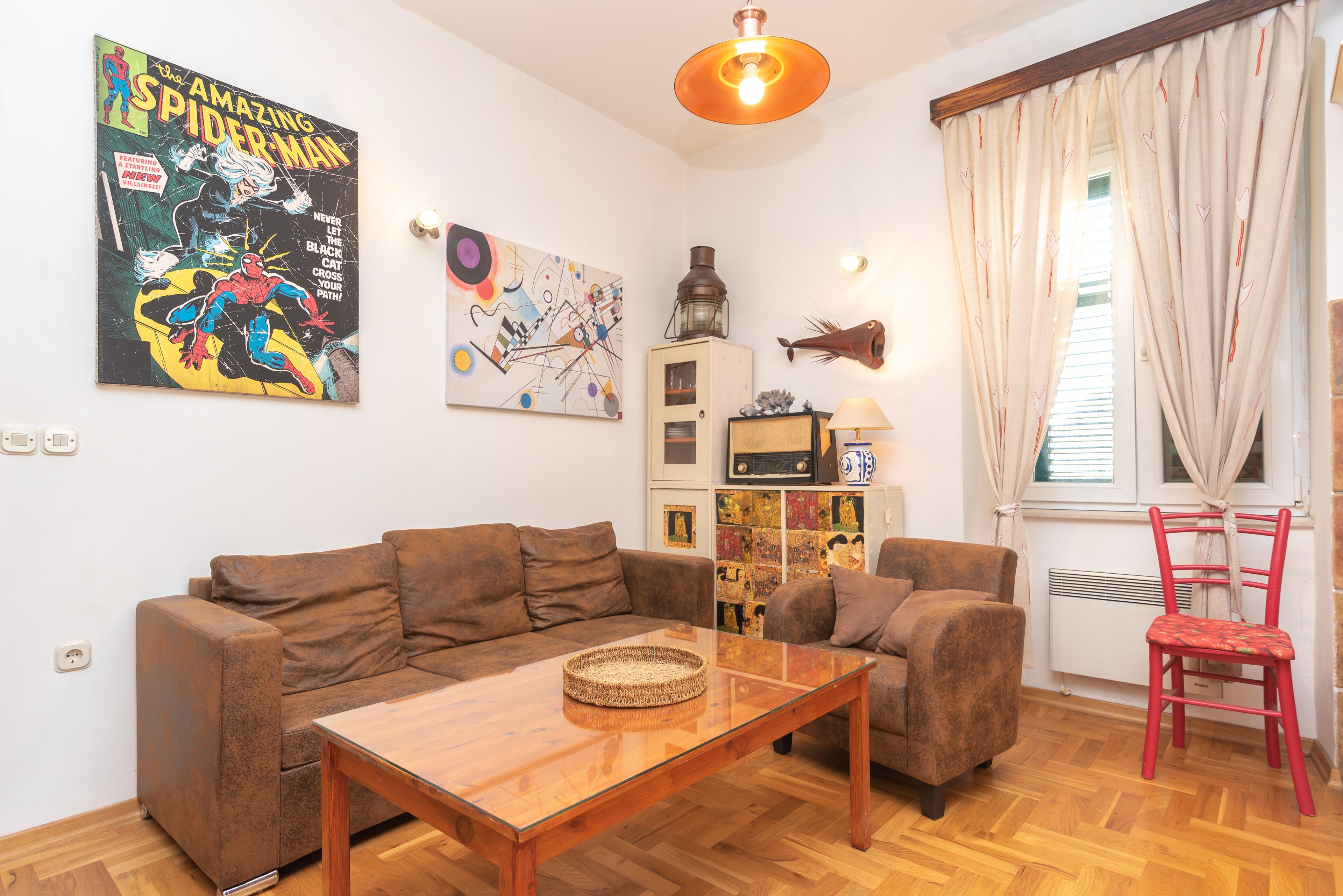 A living room with a brown sofa, a glass coffee table, and a red chair. There is a Spiderman comic on the wall, a lamp, and a fish wall decoration.