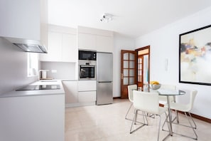 A modern kitchen with stainless steel appliances, a dining area with a glass table and white chairs, and a large abstract painting on the wall.