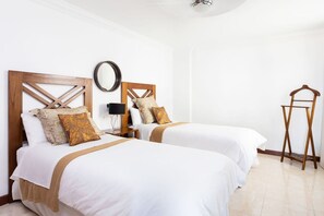 A hotel room with two beds, a wooden headboard, a round mirror, a bedside table with a lamp, and a wooden drying rack.
