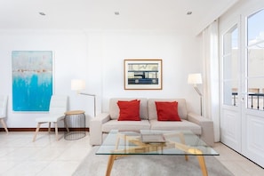 A modern living room with a glass coffee table, a sofa with red cushions, a large abstract painting, and a framed picture on the wall.