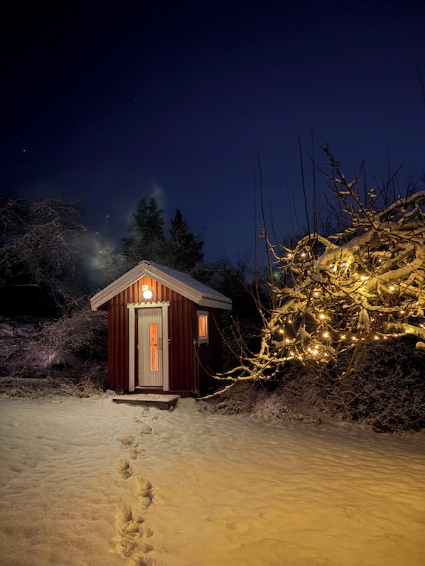A small wooden cabin with a single door and a light on the front, surrounded by snow-covered ground and trees.
