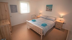 A bedroom with a single bed, two bedside tables with lamps, a window with blinds, a wall-mounted heater, and a wooden wardrobe. - Coastal Apartments - Wales (Ground Floor) (Llanelli)