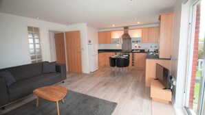 A modern living room with a grey sofa, a wooden coffee table, and a small rug. There is a kitchen area with wooden cabinets, a dining table with chairs, and a TV. - Coastal Apartments - Wales (Ground Floor) (Llanelli)