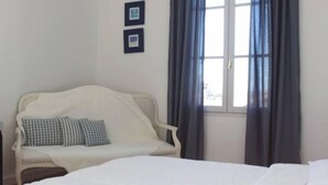 A bedroom with a bed, a white armchair with a checkered pillow, a window with curtains, and two framed pictures on the wall. - Beautiful and quiet apartment in the heart of the Cité des Papes (Avignon)