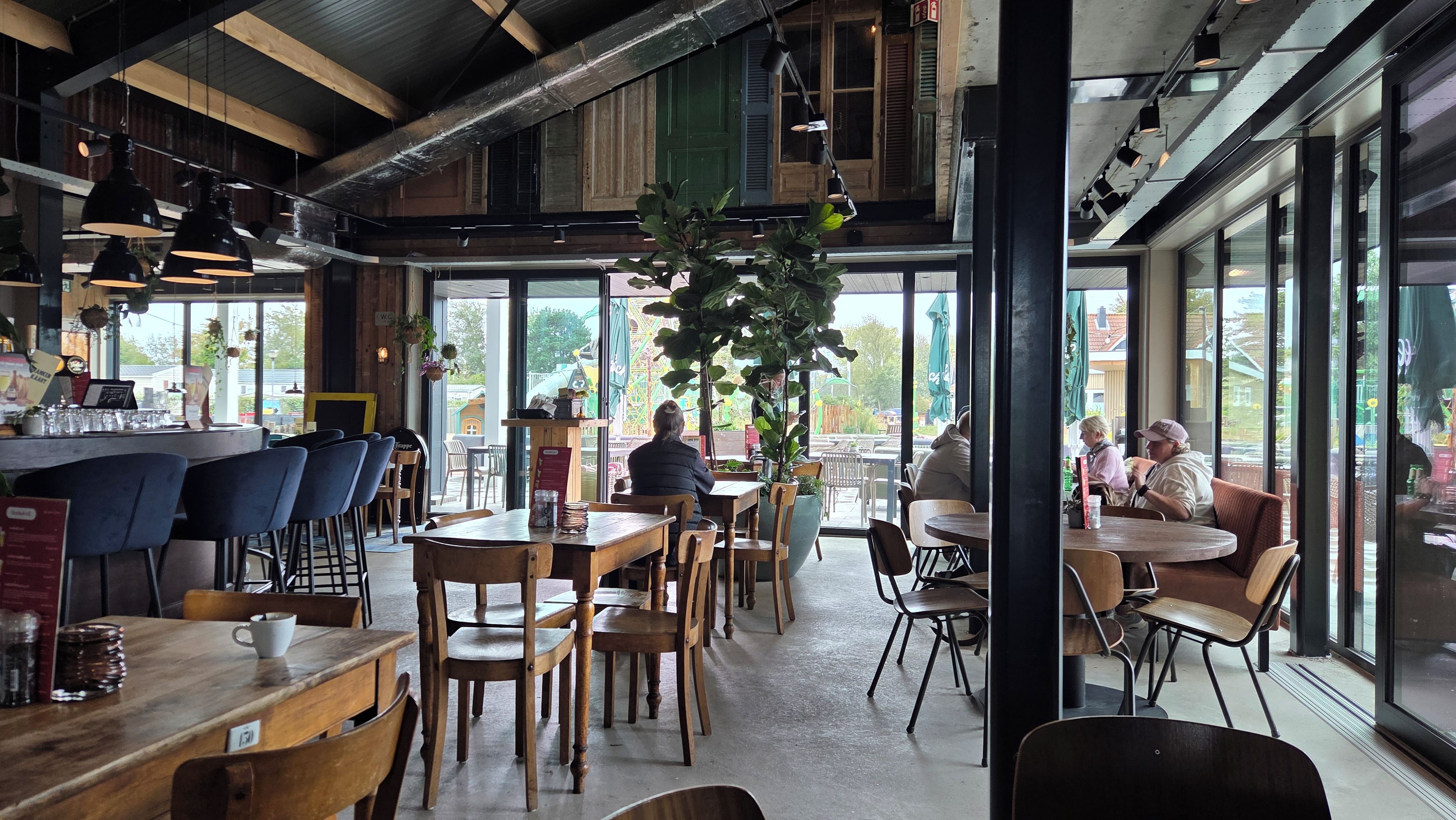 En caféindretning med træborde og stole, en bardisk og store vinduer med udsigt ud