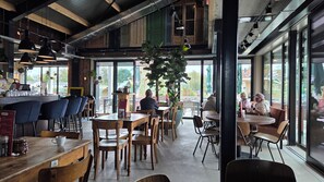 L’intérieur d’un café, avec des tables et des chaises en bois, un comptoir et de grandes fenêtres donnant sur l’extérieur.