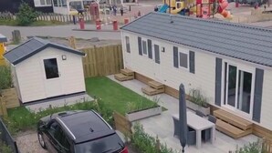A mobile home with a car parked in front, a patio area, and a playground in the background.