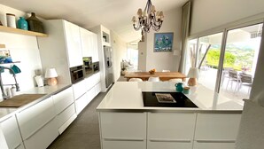 A modern kitchen with white cabinetry, a central island, and a dining area with a wooden table and chairs. - Breathtaking sea view in exclusive design villa!  (Tourrettes-Sur-Loup)