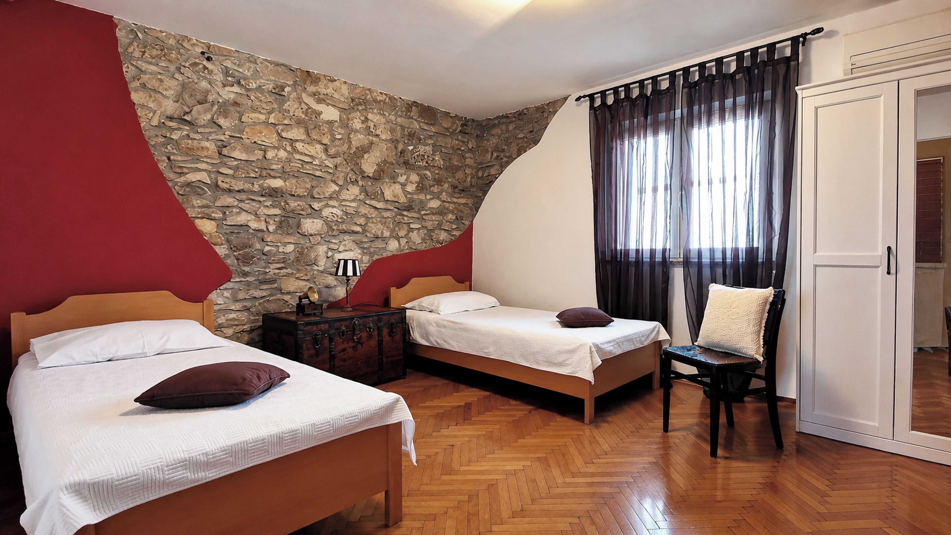 A bedroom with two beds, a stone wall, a wooden floor, and a window with curtains.