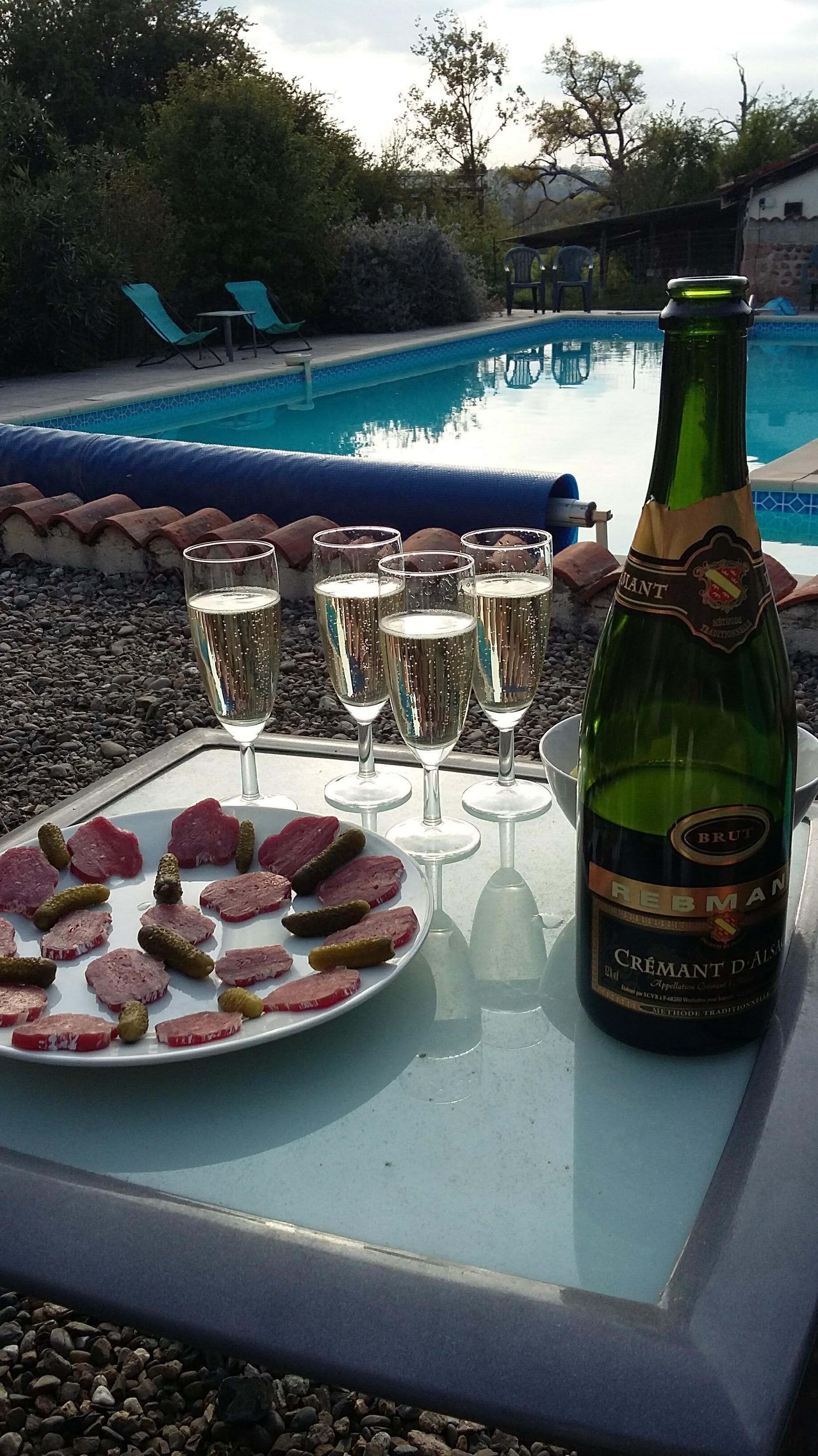 A table with a bottle of champagne, glasses, and a plate of sliced meats and pickles by a pool.